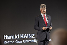 Mann mit dunklem Anzug und roter Krawatte steht auf einer Bühne und spricht. 