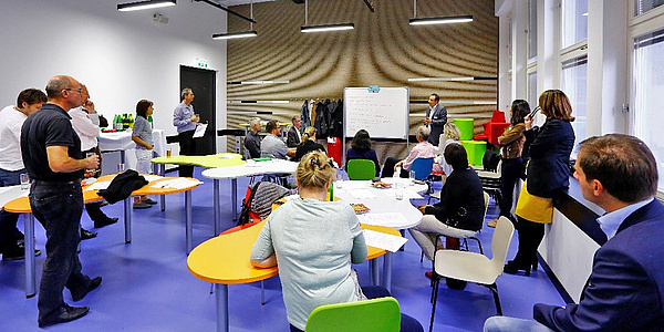 Several people are sitting and standing in a room. In the background, a man is standing next to a whiteboard.