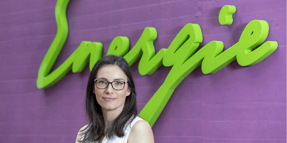 Woman with glasses and long dark hair stands in front of a green lettering with energy written on it.
