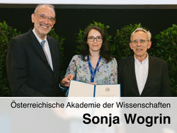 Drei Personen stehen bei einer feierlichen Übergabe vor grünen Pflanzen, die mittlere Frau hält eine Urkunde, darunter steht der Text „Österreichische Akademie der Wissenschaften – Sonja Wogrin“.