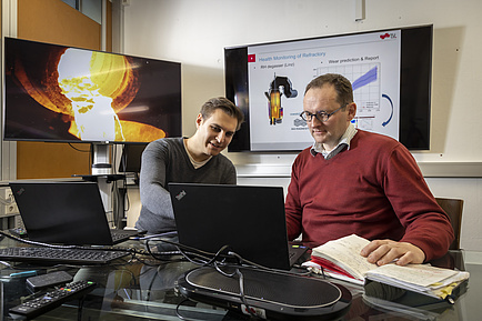 Two men talk to each other and look at a laptop