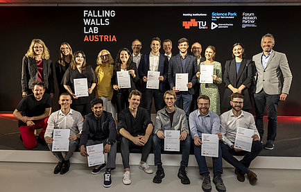 A group of people is smiling into the camera, behind them is a black wall with Falling Walls Lab Austria written on it