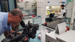 In front a man gets an apple on his camera, behind a scientific woman undertakes microbiological work