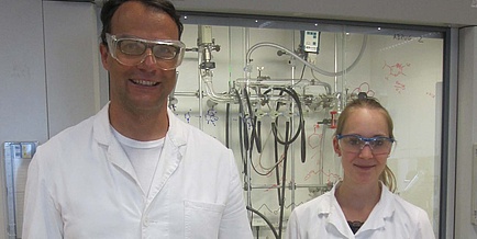 A man and a woman standing next to each other in a chemical laboratory wearing a white coat and goggles