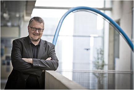 A man leans against a concrete railing and smiles into the camera.