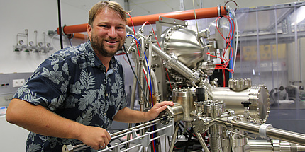 Florian Lackner. He wears short blond hair and a blue shirt with blossoms. He stands infront of the photoemission electron microscope.