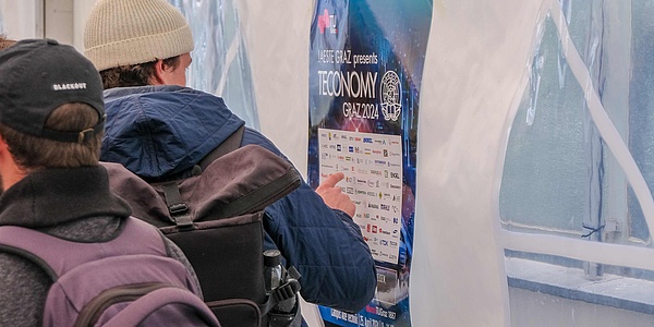 Two men with backpacks are standing with their backs to the viewer in front of an advertising poster with the words ‘IAESTE Graz presents TECONOMY Graz 2024’ and many logos.