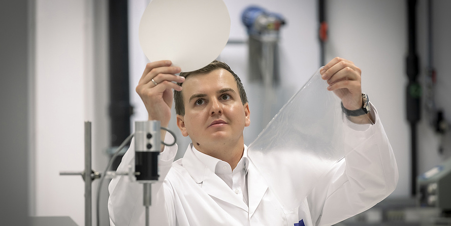 Samir Kopacic stands in his office wearing a white lab coat. He is holding a piece of paper in the one hand and a piece of plastic in the other.