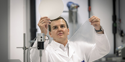 Samir Kopacic steht mit einem weißen Labormantel im Büro. In einer Hand hält er ein rundes, weißes Stück Papier, in der anderen ein Stück Plastikfolie.