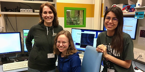 Three women in a laboratory. Screens and various measuring systems can be seen behind them.