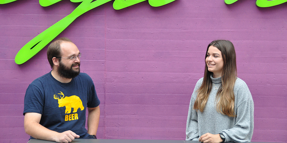 Mann mit Bart und junge Frau vor violetter Wand, an einem Tisch sitzend im Gespräch