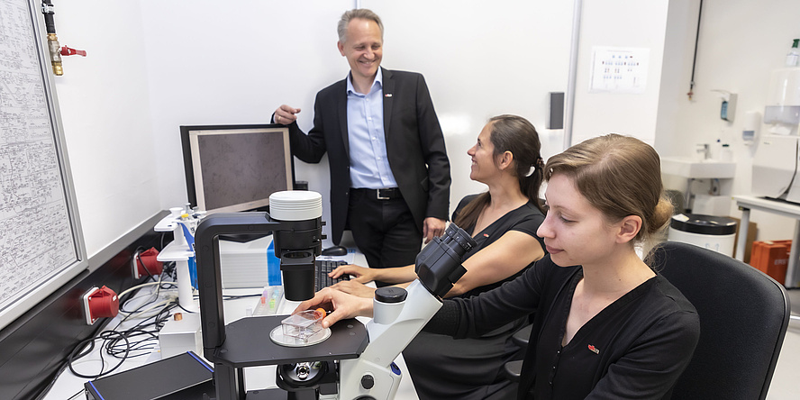 Three people at a desk which contains a microscope and a computer.