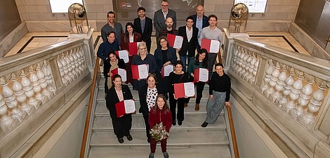Eighteen people dressed in festive attire and carrying red document folders stand on a ceremonial staircase.