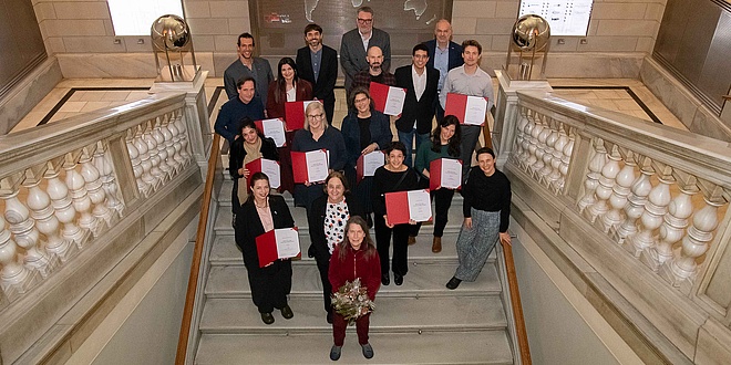 Eighteen people dressed in festive attire and carrying red document folders stand on a ceremonial staircase.