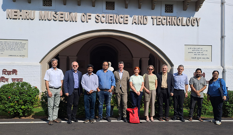 11 Personen stehen vor einem Universitätsgebäude. Das Foto ist in Indien gemacht, da indische Schriftzeichen auf dem Gebäude zu sehen sind 
