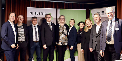 Zehn Personen stehen in einer Reihe vor einer Posterwand und schauen in die Kamera.
