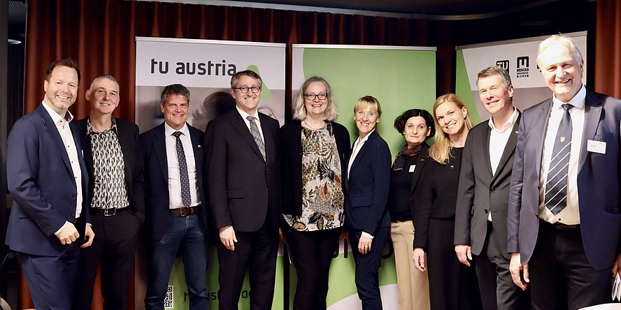 Zehn Personen stehen in einer Reihe vor einer Posterwand und schauen in die Kamera.