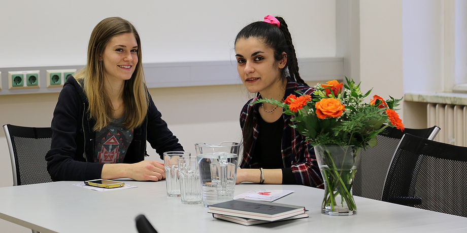 Betreuerin Nina, blond und langhaarig, mit Buddy Maria, die dunklen Haare zu einem hohen Zopf gebunden, beim Interview an einem weißen Tisch mit Notizbüchern, Wassergläsern und einem orangen Blumenstrauß.