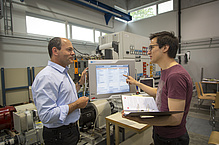 Two men in front of a control screen, electrotechnical equipment behind it.