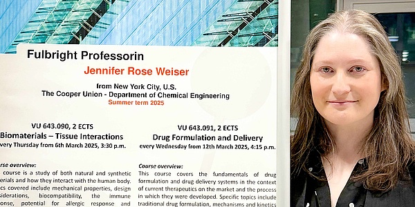 Eine Frau mit langen, dunklen, grau-melierten Haaren steht neben einem Plakat. Auf dem Plakat finden sich Ankündigungen zu den Vorlesungen der Frau. 