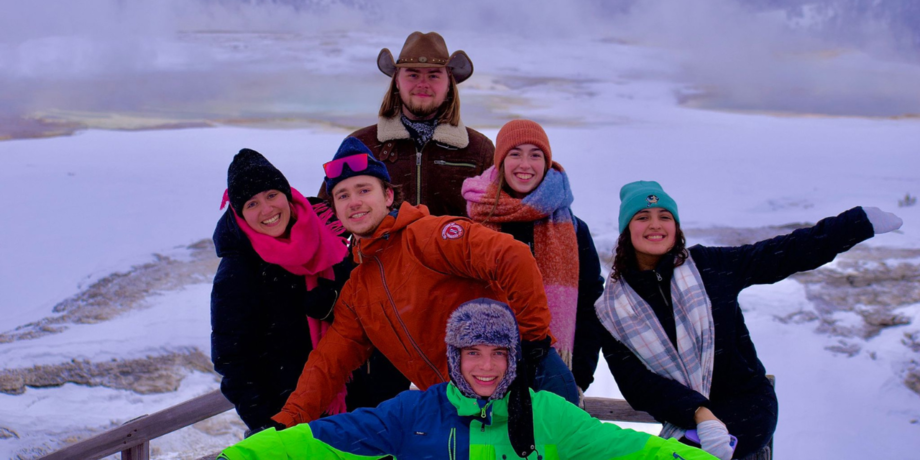 Eine Gruppe junger Menschen in bunten Skianzügen steht vor einer verschneiten Landschaft. 