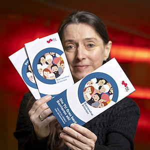 Eine Frau hält drei Flyer in die Kamera. Auf einem steht: Die TU Graz baut Barrieren ab!