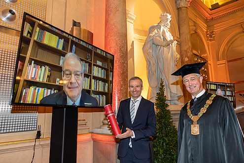 Two festively dressed gentlemen, one with a scroll of certificates, another gentleman is watching via TV.