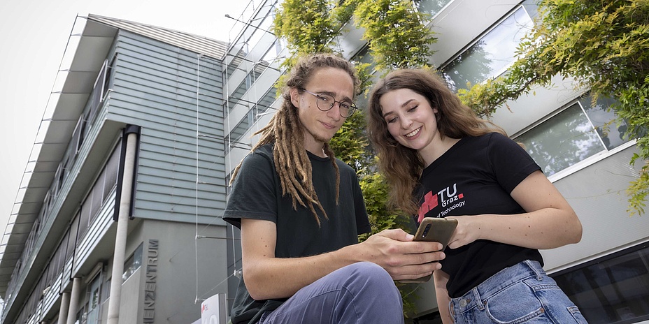 A young man and a young woman in front of a modern building that is partially overgrown with foliage.