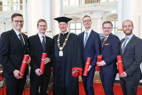 Six men smile into the camera. The third from the left wears a festive robe, the other five hold red document rolls.