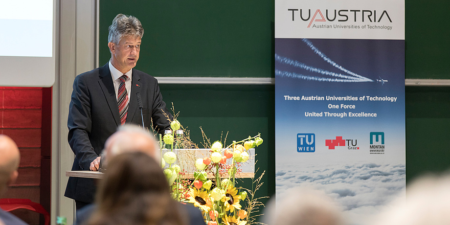 Ein Mann steht an einem Podium und spricht zum Publikum. Links von ihm steht ein Roll-Up der TU Austria. Im Vordergrund sind die Hinterköpfe der Zuhörenden zu sehen.