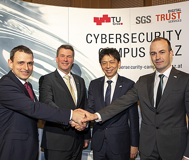 Four men in suits stand in front of the expo wall to the Cybersecurity Campus Graz and shake hands with each other.