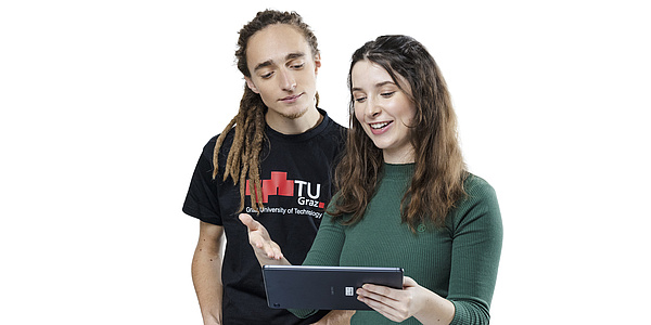 Ein junger Mann in schwarzem T-Shirt und eine junge Frau in grünem Pullover blicken gemeinsam auf ein Tablet. Die junge Frau hält das Tablet in der linken Hand, mit der rechten deutet sie darauf.