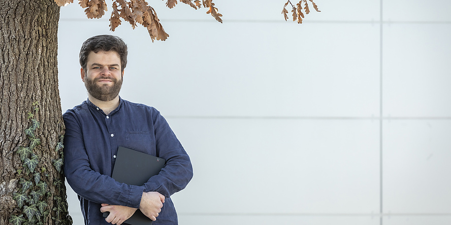 David Garcia steht an einen Baum gelehnt und hält seinen Laptop in der Hand.