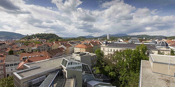 Blick über die Dächer des historischen Zentrums einer Stadt mit einem modernen Gebäude im Vordergrund, blauer Himmel mit malerischen Wolken.