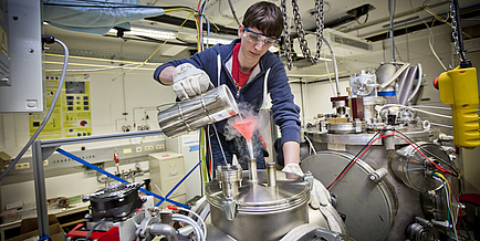 Junger Mann mit blauer Sweatjacke und Schutzbrille schüttet in einem physikalischen Labor eine Flüssigkeit durch einen roten Trichter.