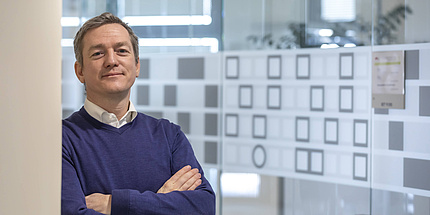 Portrait of a man in a blue jumper and white shirt, leaning against a column with folded arms