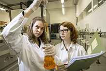 Zwei Frauen in Laborkitteln entnehmen mit einer Pipette eine Flüssigkeit aus einem kolbenförmigen Glasgefäß.
