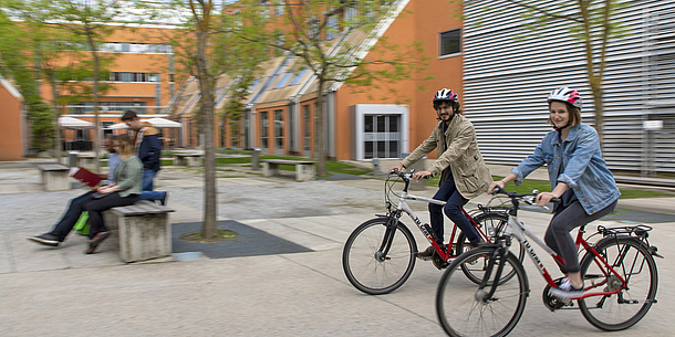 Auf einem Platz mit einigen Bäumen fahren eine Studentin und ein Student mit TU Graz-Rädern. Im Hintergrund sind Studierende auf einer Bank zu sehen.