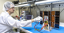 A man in a lab coat and protective hood investigates a wired satellite.