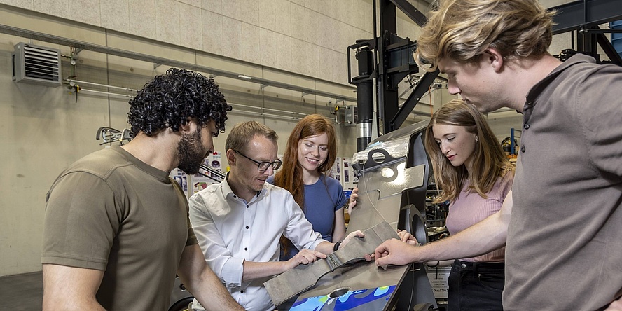 Ein Lehrender erklärt vier umstehenden Studierenden in einem Großraumlabor ein Werkstück aus Metall.