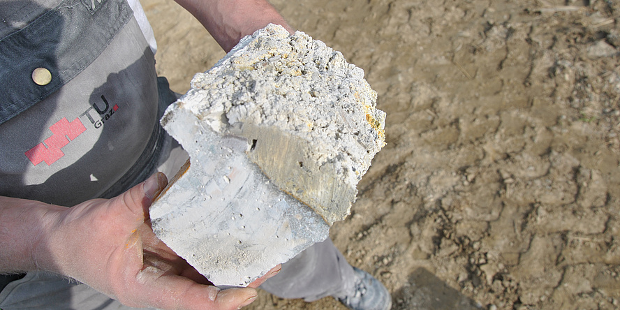 A male hand holds a concrete part damaged by microbial induced concrete corrosion (MICC); a centimetre-thick deposit has formed on the part.