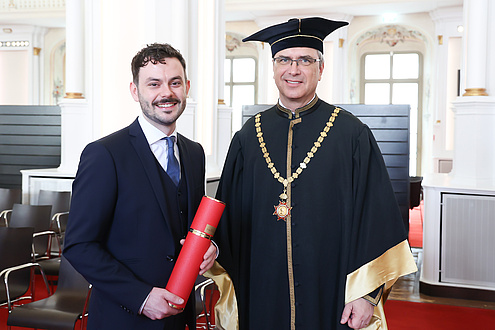 [Translate to Englisch:] Two men smile into the camera. The man on the left is holding a red document roll and the man on the right is wearing a festive robe.