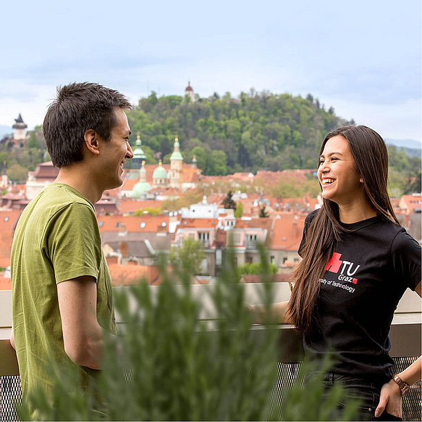 Ein Mann und eine Frau unterhalten sich auf einem Balkon. Im Hintergrund die Stadt Graz. Bildquelle: Lunghammer – TU Graz