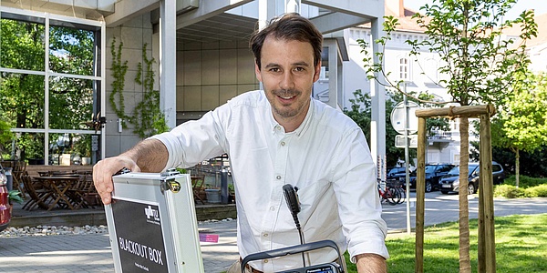 Ein Mann mit braunen Haaren, Dreitagebart und weißem Hemd lächelt in die Kamera. Dabei hat er einen Koffer geöffnet. Auf dem Koffer steht "Blackout Box".