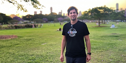 Ein Mann mit dunklen Haaren und einem schwarzen T-Shirt schaut in die Kamera. Er steht in einem Park, im Hintergrund sind Hochhäuser zu sehen. 