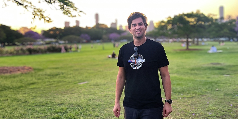 A man with dark hair and a black T-shirt looks into the camera. He is standing in a park, with skyscrapers in the background.