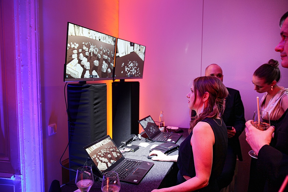 Eine Dame im Ballkleid steht vor zwei Bildschirmen und versucht anhand von Virtual Reality einen Mauer zu bauen.