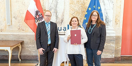 Ein Mann und zwei Frauen vor der Österreich-Fahne, eine Dame hält eine Urkunde in Händen