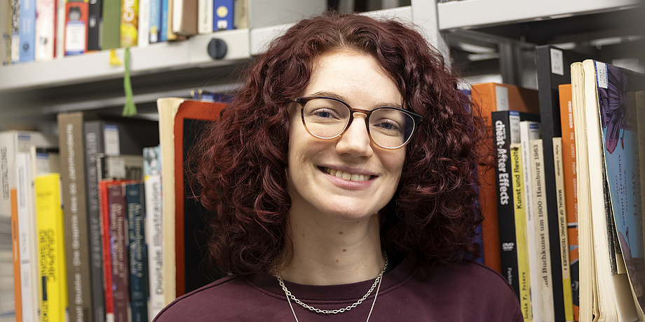 A woman with glasses smiles into the camera, many books can be seen in the background.