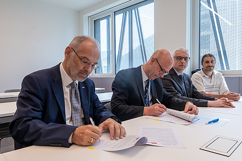 Vier Männer sitzen an einem Tisch. Die beiden auf der linken Seite unterzeichnen gerade ein Schriftstück, die zwei daneben sehen dabei zu.